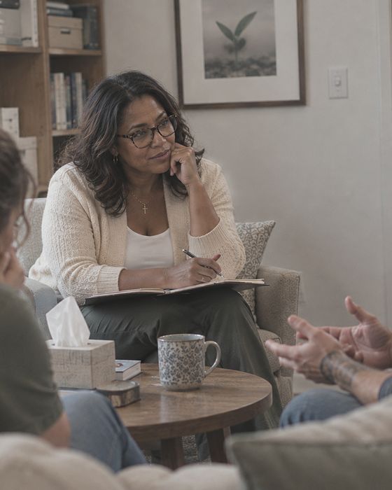 A therapist listens thoughtfully, notebook in hand, as a couple talks with her in a calm, lived-in office.